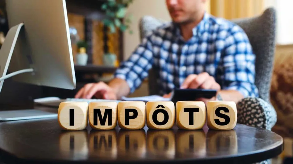 Des dés en bois épellant le mot français "IMPÔTS" (taxes) devant un homme travaillant sur un ordinateur et une calculatrice.