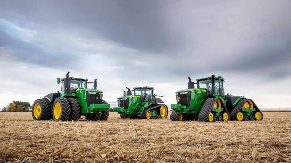 Trois tracteurs John Deere verts et jaunes dans un champ labouré sous un ciel nuageux.