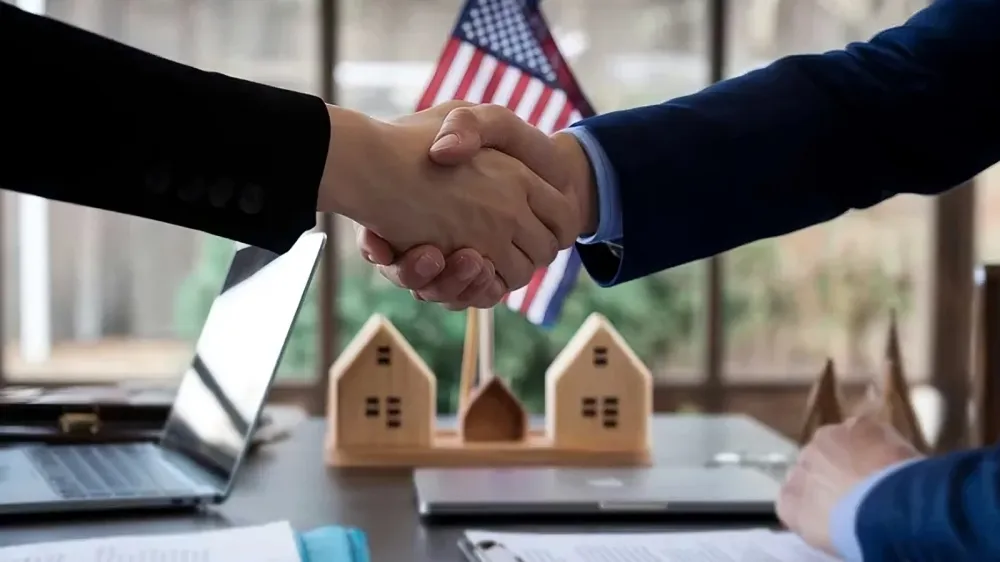 Poignée de main symbolisant un accord immobilier, avec un drapeau américain en arrière-plan et des maquettes de maisons sur la table.